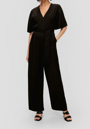 Kvinde iført en sort jumpsuit i wrap-stil med korte ærmer, brede ben, et bundet bælte i taljen og sorte sandaler med åben tå.