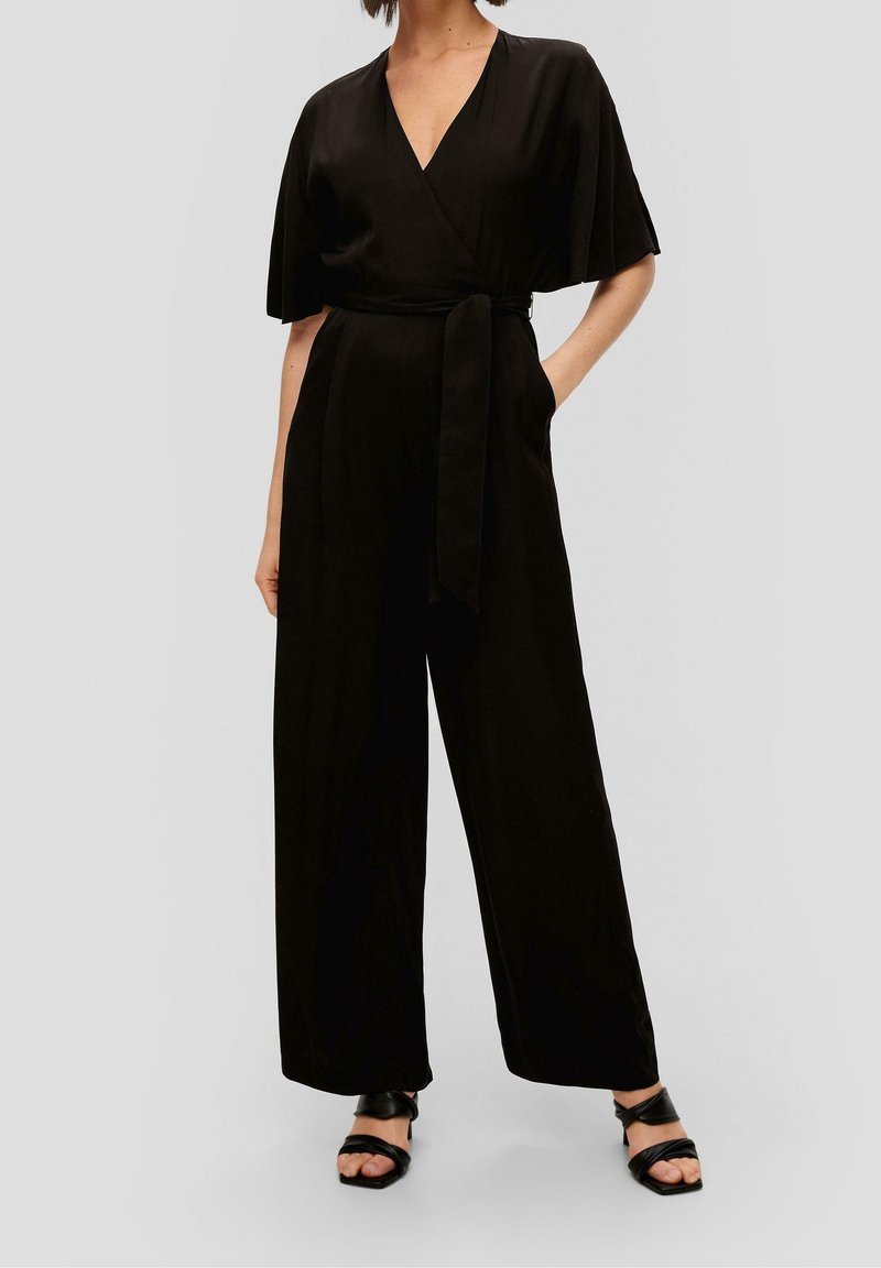 Femme portant une combinaison noire style cache-cœur avec manches courtes, jambes larges, ceinture nouée à la taille et sandales noires à bouts ouverts.