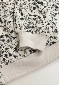 Vaaleabeige kangas mustalla kukkaompeleella ja pienillä helmillä, esittäen pitkähihaisen vaatteen resorikärkeä ja vyötäröosaa.