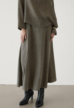 Femme portant une longue jupe marron, un haut marron assorti, des bottes à talons noires et un bracelet en or sur un fond gris clair.