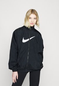 Črn najlon jopič z visokim ovratnikom, sprednjim zadrgo, elastičnimi manšeti in velikim belim Nike swooshom na prsih.