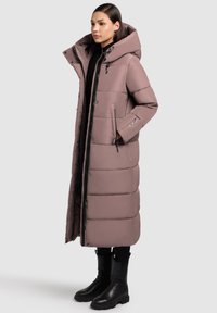 Ilga pūkinė striukė švelnios mauvės spalvos, su aukštu apykakliu, užtrauktukais užsegamomis kišenėmis ir elegantišku siuvinėtu dizainu. Derinama su juodais kulkšnies aulinukais.