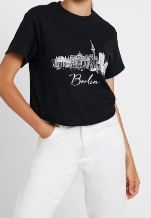 Musta puuvillainen T-paita, jossa valkoinen graafinen kuva Berliinin siluetista ja "Berlin"-teksti tyylikkäällä fontilla. Lyhyet hihat, rento istuvuus.