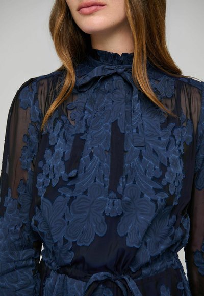 Femme portant un chemisier bleu marine transparent avec une grande broderie florale, un col haut à volants et un nœud noué au cou.