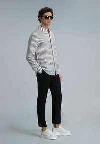 Chemise à rayures à manches longues en crème et marron, pantalon noir et baskets blanches. Modèle portant des lunettes de soleil noires, se tenant devant un fond neutre.