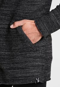 Maglione testurizzato nero e grigio con tasca laterale e cuciture visibili, con un'etichetta sul lato sinistro in basso.