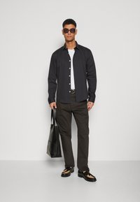 Camicia nera a bottone sopra una maglietta bianca, abbinata a pantaloni marroni scuri e scarpe nere. Una borsa a tracolla nera e occhiali da sole completano il look.
