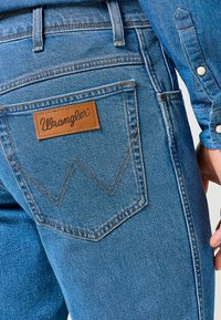 Vaqueros de mezclilla azul con una etiqueta Wrangler en el bolsillo trasero, que cuentan con costuras clásicas y una ligera textura. Diseño estándar de cinco bolsillos.