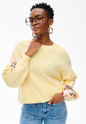 Kobieta o krótkich włosach nosi duże okulary, złote kolczyki koła z koralikami, żółty sweter z kwiatowymi mankietami oraz niebieskie dżinsy, pozując pewnie.