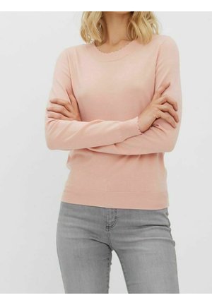 Femme aux bras croisés portant un pull rose clair à bords festonnés et un jean gris sur un fond uni.