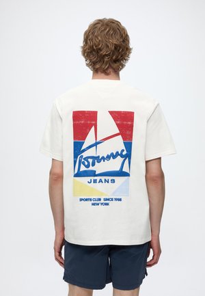 Personne aux cheveux blonds portant un t-shirt blanc avec un graphisme rouge, bleu et jaune d’un voilier et le texte « Tommy Jeans Sports Club Since 1985 New York »