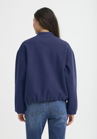 Tumši zila, pārspīlēti liela sporta krekls ar augstu apkakli, kritušām plecu šuvēm un ribotām maliņām, izgatavots no mīksta, teksturēta auduma.