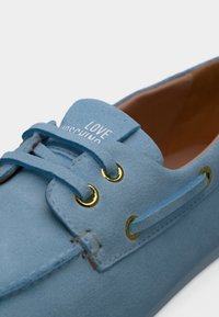 Γαλάζιο μοκασίνι από σουέτ με χρυσά ρούμπανια και κορδόνια, με το κείμενο "LOVE MOSCHINO" στη γλώσσα.