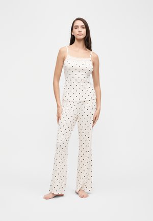 Une femme porte un caraco blanc et un pantalon de pyjama assorti à jambes larges et à pois noirs, sur un fond uni.