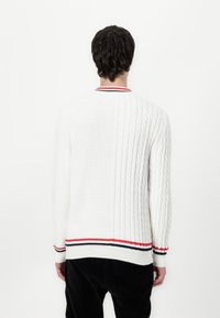 Weißer Kabelstrickpullover mit strukturiertem Rücken, verziert mit roten und schwarzen Streifen am Kragen und Saum. Lange Ärmeln.