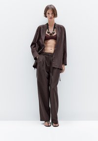 Bruine broek met fijne strepen, losvallend, met een bijpassende open jas, gedragen over een bordeauxrode top. Opvallende ronde zwarte halsketting en sandalen.