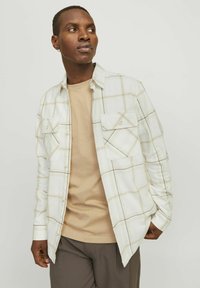 Camisa de cuadros blanca con líneas beige y marrones, dos bolsillos en el pecho, diseño de botones, llevada sobre una camiseta beige y pantalones marrones.