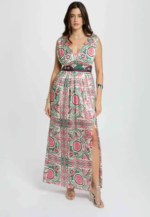 Donna con abito senza maniche, lungo fino al pavimento, con motivi floreali rosa e verdi, spacco laterale e sandali argento con tacco alto su sfondo neutro.