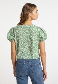 Grüne Bluse mit Blumenmuster, Puffärmeln und einem Schlüsselaugenverschluss im Rücken, kombiniert mit hoch taillierten blauen Jeans. Baumwollstoff mit strukturiertem Muster.