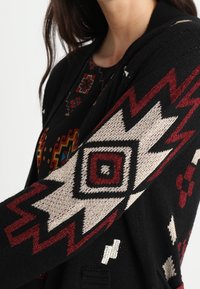 Cardigan nero con un motivo geometrico in rosso e crema sulle maniche, con una morbida texture in maglia e un design aperto sul davanti.