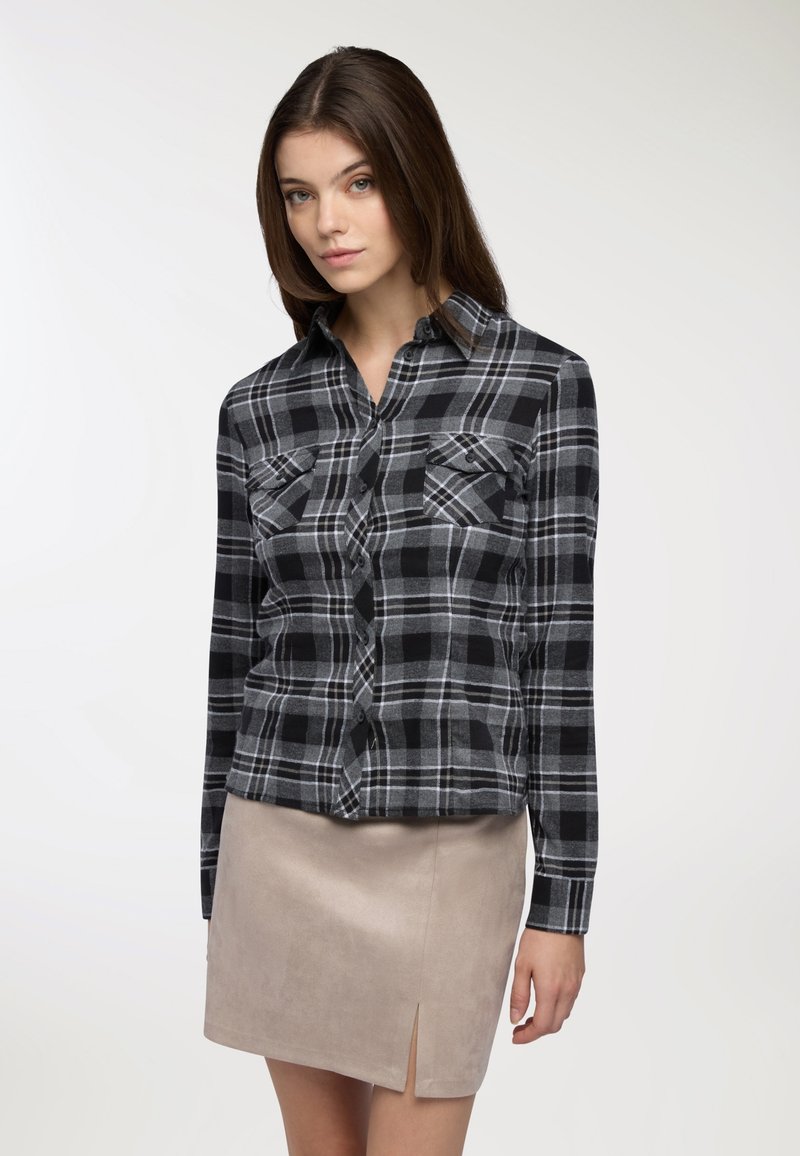 Camicia di flanella a quadri neri e grigi con due tasche sul petto, chiusura con bottoni e maniche lunghe, abbinata a una gonna in suede beige chiaro con uno spacco.