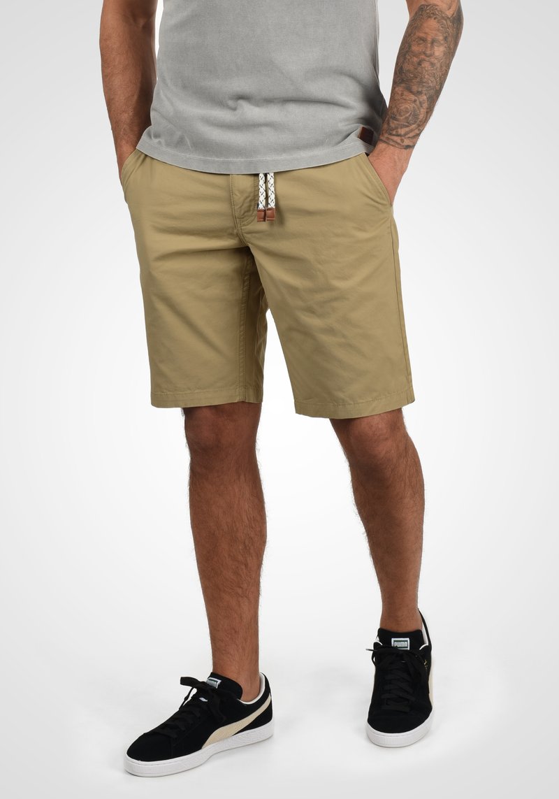 Pantalones cortos caqui con un frente plano, dos bolsillos laterales y una cintura con cordón en negro y blanco. Combinados con zapatillas negras.
