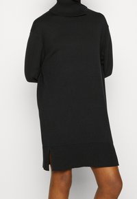 Schwarzes gestricktes Rollkragenkleid mit gerippten Abschlüssen und seitlichen Schlitzen. Hat eine lockere Passform und lange Ärmel. Glatte Textur, knielanges Design.