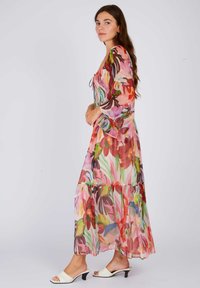 Floral maxi dress in sheer fabric with vibrant pink, green, and yellow patterns. Features a smocked bodice and flared sleeves, worn with white mules.