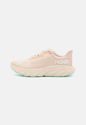 Chaussure de course Hoka beige clair avec tige en mesh respirant, semelle épaisse amortie, fermeture à lacets, et accents de semelle extérieure bleu aqua.