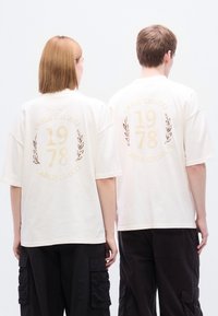 Τα υπερμεγέθη μπλουζάκια σε κρεμ χρώμα διαθέτουν ένα μεγάλο τυπωμένο σχέδιο με τη φράση «CARLO COLUCCI», την ένδειξη του έτους «1978» και διακοσμητικές δάφνινες.