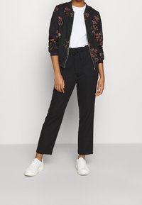 Veste bomber noire avec broderie florale, associée à un t-shirt blanc et un pantalon noir. Le mannequin porte des baskets blanches, debout avec assurance.