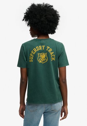 Persoon met krullend haar, van achteren gezien, draagt een groen T-shirt met gele "Superdry Track" tekst en embleem, gecombineerd met lichtblauwe jeans.