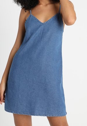 Femme portant une robe en denim bleu sans manches avec de fines bretelles et un décolleté en V, posant avec un bras levé et l'autre détendu.