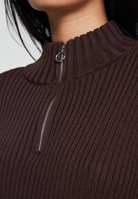 Brauner gerippter Pullover mit hohem Kragen und silbernem Reißverschlussdetail. Der strukturierte Stoff verleiht ein gefügiges Aussehen und betont den Ausschnitt.