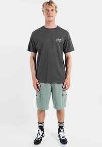 T-shirt en coton gris avec petit logo blanc sur la poitrine, accompagné d'un short cargo vert clair. Baskets montantes noires avec chaussettes rayées.