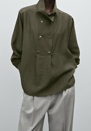 Osoba ubrana w luźną oliwkowozieloną bluzę na guziki z wysokim kołnierzem i jasnoszare spodnie w szerokie szare prążki, ręce w kieszeniach.