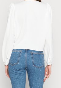 Femme portant un chemisier blanc à manches longues et un jean denim bleu à petits pois blancs, vue de dos.