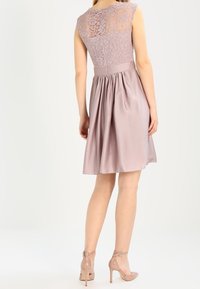 Robe sans manches à dos en dentelle, en mauve doux, avec un corsage ajusté et une jupe évasée. Texture lisse avec une finition satinée, accompagnée de talons nus.