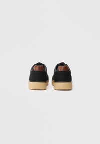Sneakers neri e marroni con suole beige mostrati da dietro su uno sfondo grigio chiaro, centrati e uniformemente distanziati.