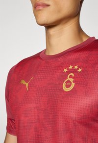 Piros sport póló textúrált mintával, arany logóval és kiegészítőkkel. Kerek nyakkivágással és rövid ujjakkal rendelkezik.