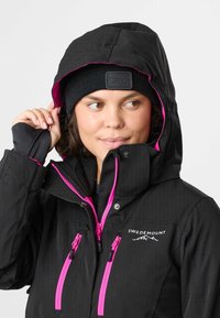 Black outdoor jacket with pink zippers and accents, featuring a hood and zippered pockets. Worn with a black beanie.