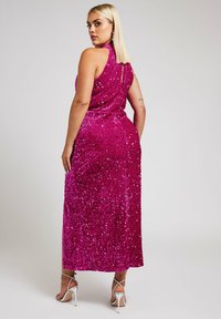 Lång, ärmlös klänning i starkt rosa med paljettmönster, hög krage och detalj med nyckelhål i ryggen. Bärs med silverfärgade högklackade sandaler.