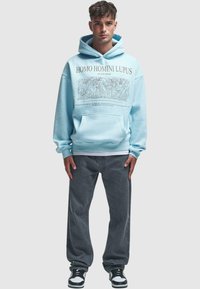 Light blue hoodie with front pocket, graphic text and illustration, paired with gray pants and black and white sneakers. Casual fit.