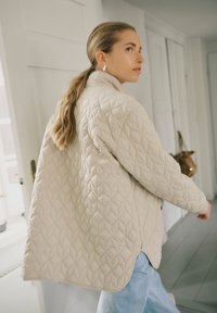 Pikkade juustega naine hobusesabas, kandes beežikat kapuutsiga jopet ja siniseid teksaseid, kõnnib siseruumides heledas, minimalistlikus koridoris.