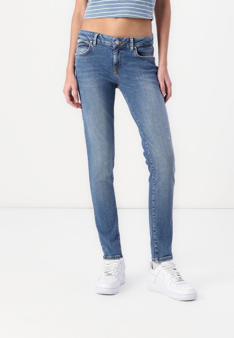 Világoskék, slim-fit farmernadrág öt zsebbel, elülső gombos zárással és enyhe fakulással. Fehér sportcipővel párosítva.