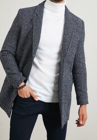 Navyblauw-wit patroon blazer gecombineerd met een witte coltrui, met een ontspannen pasvorm en een gestructureerde stof, gedragen over een donkergekleurde broek.