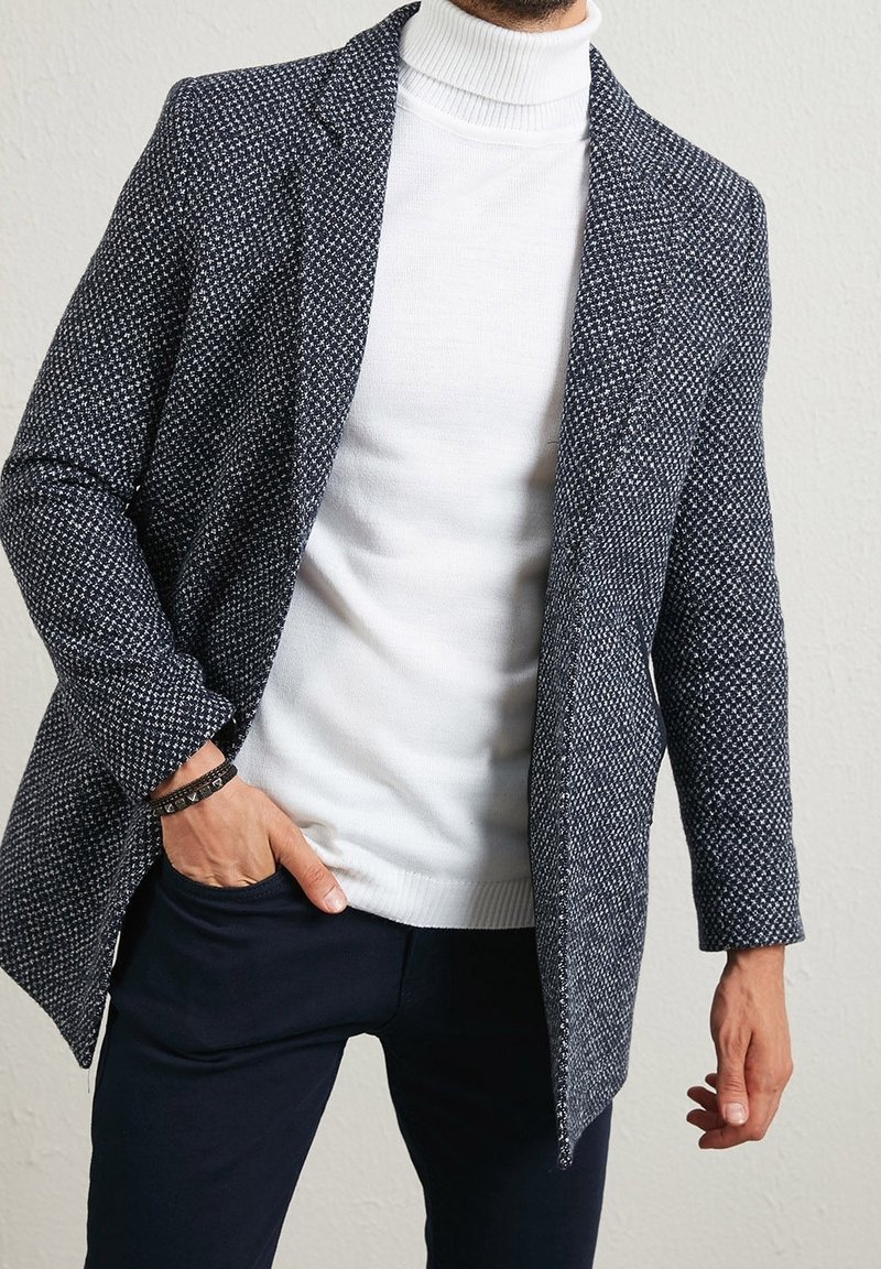 Navyblauw-wit patroon blazer gecombineerd met een witte coltrui, met een ontspannen pasvorm en een gestructureerde stof, gedragen over een donkergekleurde broek.