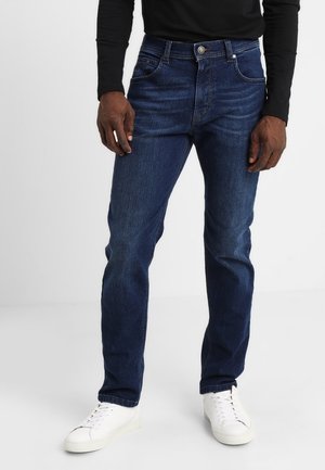 Homme portant un jean slim bleu foncé, une chemise noire à manches longues et des baskets blanches, debout sur un sol blanc avec un fond uni.