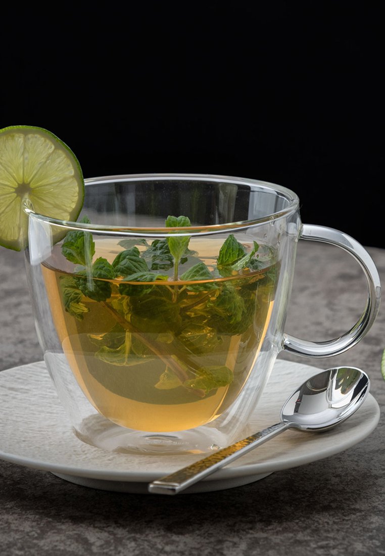 Przezroczysta szklanka z ziołową herbatą, z listkami mięty i plasterkiem limonki, na białym spodeczku, obok niej srebrna łyżeczka.