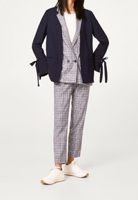 Nainen, jolla on päällä vaalean ruudullinen puku, kaksirivinen takki, tummansininen blazer, jossa on solmittavat hihadat, ja valkoiset lenkkarit, seisoo valkoista taustaa vasten.